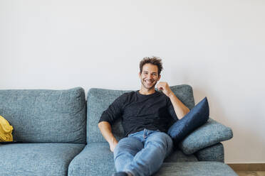 Happy young man sitting on sofa in living room at home - MEUF04717