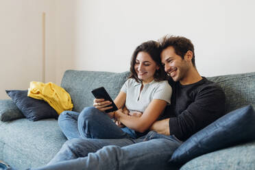 Lächelnde Freundin teilt ihr Smartphone mit ihrem Freund auf dem Sofa zu Hause - MEUF04718