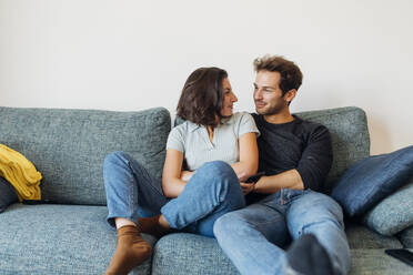 Smiling couple looking at each other while sitting on sofa - MEUF04719