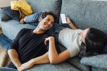 Happy young man looking at girlfriend with mobile phone on sofa at home - MEUF04721