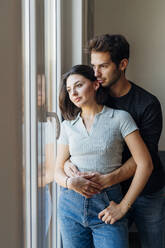 Young couple looking through window at home - MEUF04738