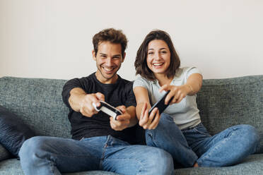 Fröhliches junges Paar spielt ein Videospiel auf dem Sofa im Wohnzimmer - MEUF04742