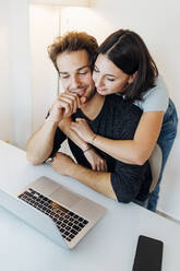 Junges Paar umarmt sich bei der Arbeit am Laptop zu Hause - MEUF04754