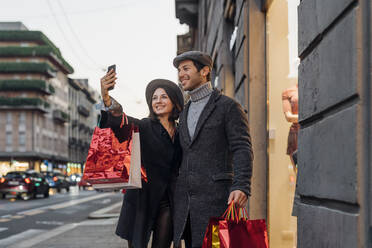 Smiling young couple with shopping bags taking selfie with smart phone near store - MEUF04767