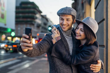 Glückliches junges Paar mit Hut nimmt Selfie durch Smartphone in der Stadt - MEUF04770