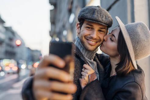 Junge Frau küsst glücklichen Freund, während sie ein Selfie auf dem Smartphone macht - MEUF04772