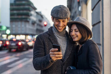 Glückliches Paar in warmer Kleidung teilt sich ein Smartphone auf der Straße - MEUF04773