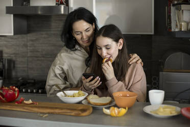 Mutter und Tochter schauen beim Frühstück in der Küche auf ihr Smartphone - LLUF00409