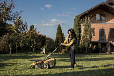 Woman mowing grass with lawn mower on sunny day - LLUF00415