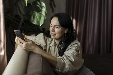 Woman using mobile phone on sofa at home - LLUF00425