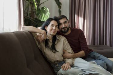 Smiling man embracing woman sitting on sofa in living room at home - LLUF00428