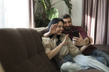 Couple waving hand to video call through mobile phone on sofa - LLUF00431