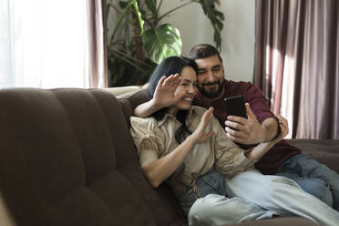 Paar winkt mit der Hand zum Videogespräch über Handy auf dem Sofa - LLUF00431