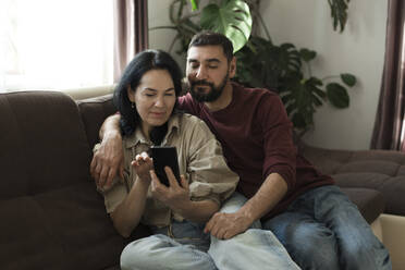 Couple using mobile phone on sofa at home - LLUF00432