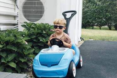 Junge mit Sonnenbrille sitzt in blauem Spielzeugauto bei Pflanzen - ACTF00150