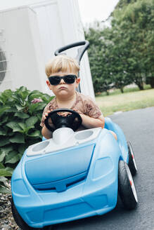 Gelangweilter blonder Junge mit Sonnenbrille, der in einem blauen Spielzeugauto sitzt - ACTF00151