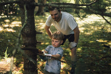 Father looking at son cutting branch of tree with hand saw - ACTF00154