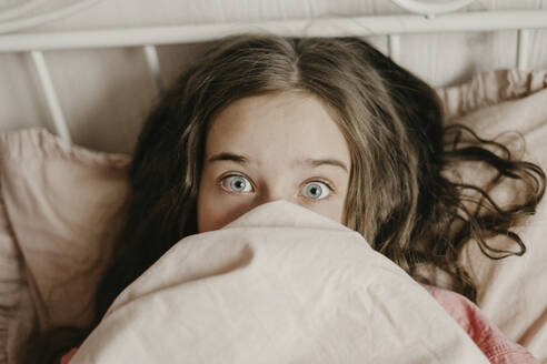 Surprised girl with gray eyes covering mouth with blanket at home - SEAF00180