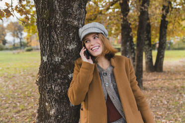 Glückliche Frau, die am Baum im Park mit ihrem Handy spricht - MGIF01114