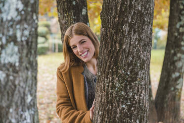 Happy young woman standing behind tree trunk - MGIF01118