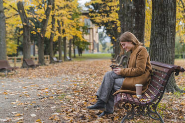 Lächelnde Frau mit Mobiltelefon auf einer Bank im Herbstpark - MGIF01122