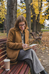 Smiling woman writing in book in autumn park - MGIF01125