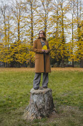 Junge Frau mit Herbstblatt auf einem Baumstumpf in einem öffentlichen Park stehend - MGIF01129