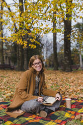 Lächelnde Frau mit Buch auf einer Decke sitzend im Herbstpark - MGIF01135