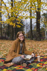 Smiling woman with book sitting on blanket in autumn park - MGIF01135