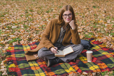 Lächelnde junge Frau sitzt im Schneidersitz auf einer Decke im Park - MGIF01136