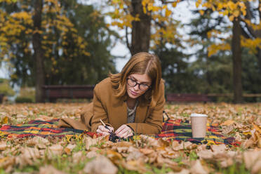 Junge Frau liegt auf einer Decke und schreibt in einem Buch in einem herbstlichen Park - MGIF01140