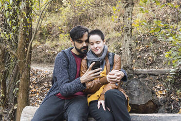 Bärtiger Mann teilt Smartphone mit Freundin auf Baumstamm am Flussufer - MGRF00580