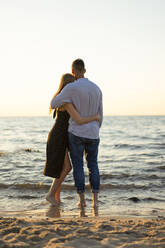 Romantic young couple with arms around looking at horizon from beach - SSGF00327