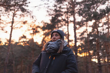 Junge Frau mit warmer Kleidung im Wald bei Sonnenuntergang - MRRF01700