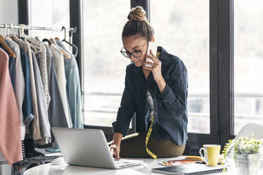 Lächelnde Geschäftsfrau, die einen Laptop benutzt und mit einem Mobiltelefon im Büro spricht - JSRF01708