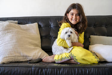 Lächelndes Mädchen sitzt mit Hund auf dem Sofa zu Hause - DLTSF02416