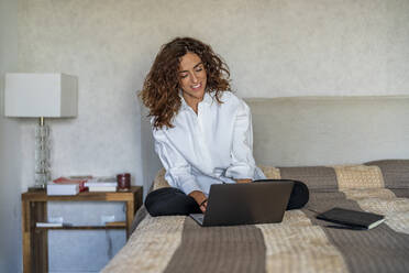 Smiling woman using laptop on bed in bedroom at home - DLTSF02423