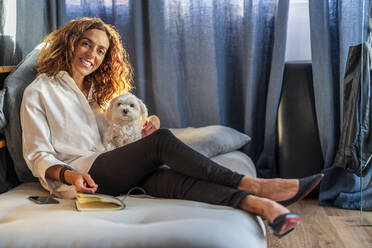 Happy woman sitting with dog on sofa in living room - DLTSF02426