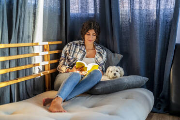 Girl reading book and sitting with dog on sofa - DLTSF02427