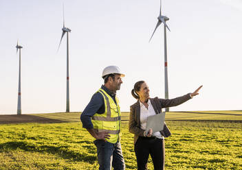 Lächelnder Ingenieur mit digitalem Tablet, der einem Kollegen im Windpark etwas erklärt - UUF25076