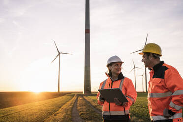 Ingenieur mit Tablet-PC lächelt und diskutiert mit einem Kollegen im Windpark - UUF25096