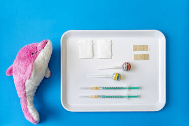 Gefülltes Delphinspielzeug neben einem Tablett mit Impfstoffinjektionen und Lutscher auf blauem Hintergrund - DRF01777