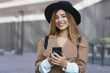 Woman wearing hat and trenchcoat holding smart phone in city - PNAF02570
