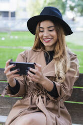 Lächelnde Frau, die auf einer Bank auf ihr Mobiltelefon schaut - PNAF02588