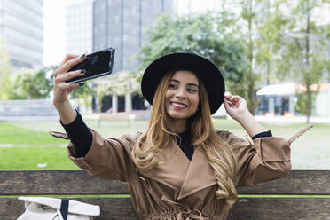 Woman wearing hat taking selfie through smart phone on bench - PNAF02591