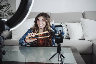 Lächelnde Friseurin, die ihr Haar mit einer Maschine lockt und über ihr Smartphone im Heimstudio vloggt - EBBF04881