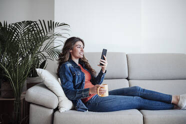 Smiling young woman using smart phone on sofa at home - EBBF04885