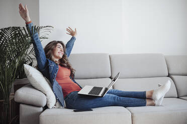 Carefree woman with laptop sitting on sofa at home - EBBF04889