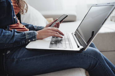 Woman with mobile phone using laptop in living room - EBBF04893