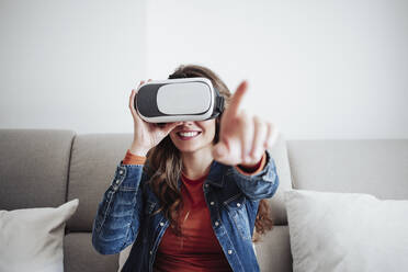 Lächelnde junge Frau mit Virtual-Reality-Headset und Gesten auf dem Sofa - EBBF04897